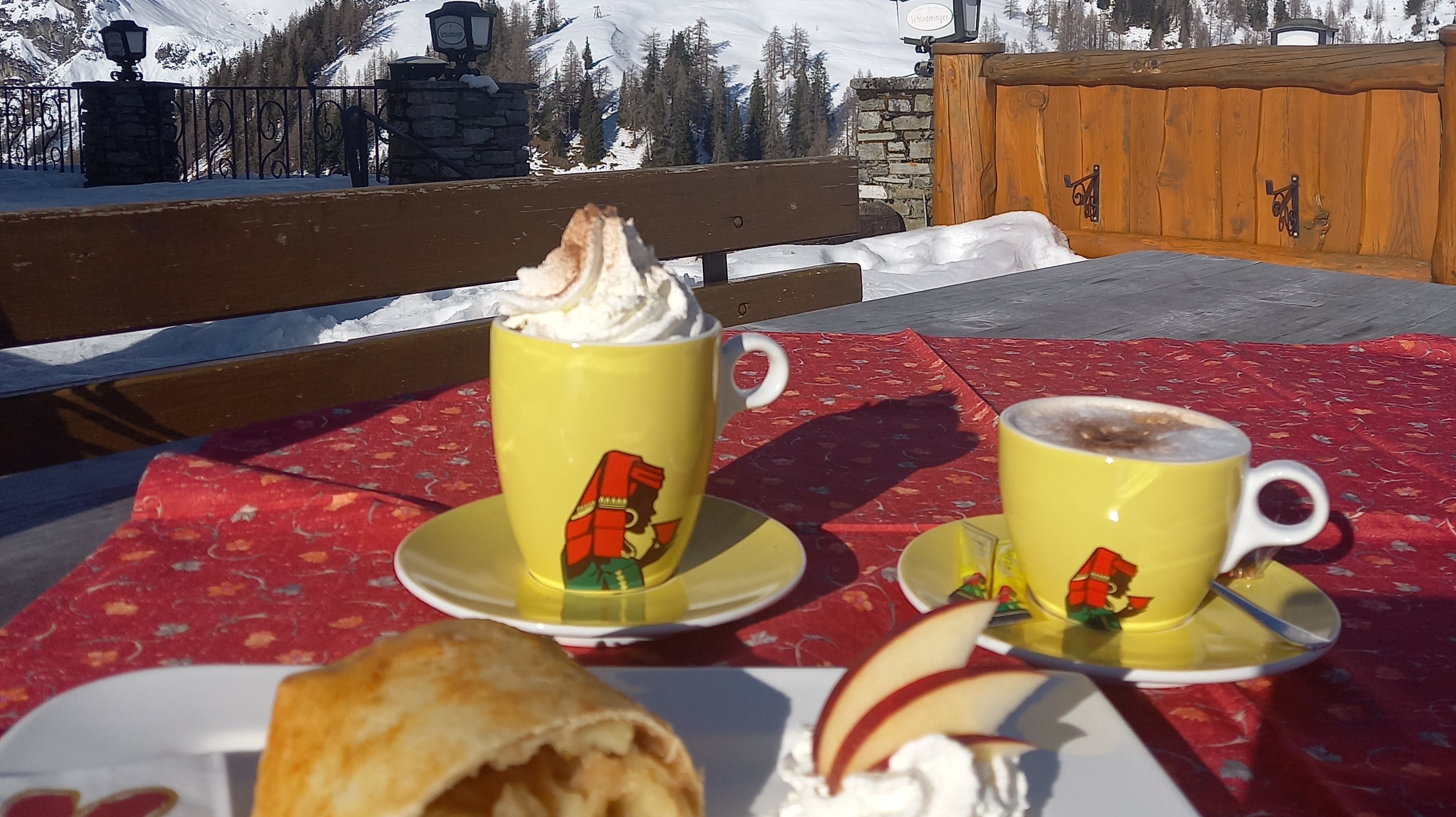 Winterliche Genüsse im Bergpanorama Ein Teller Apfelstrudel mit Sahne und Apfelscheibe, zwei Tassen heißer Getränke vor schneebedeckten Bergen.