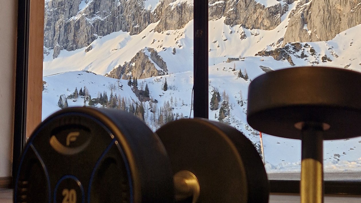 Aktiv im Schnee mit Hanteln Zwei Hanteln auf einer Matte vor einem großen Fenster mit schneebedeckten Bergen im Hintergrund.