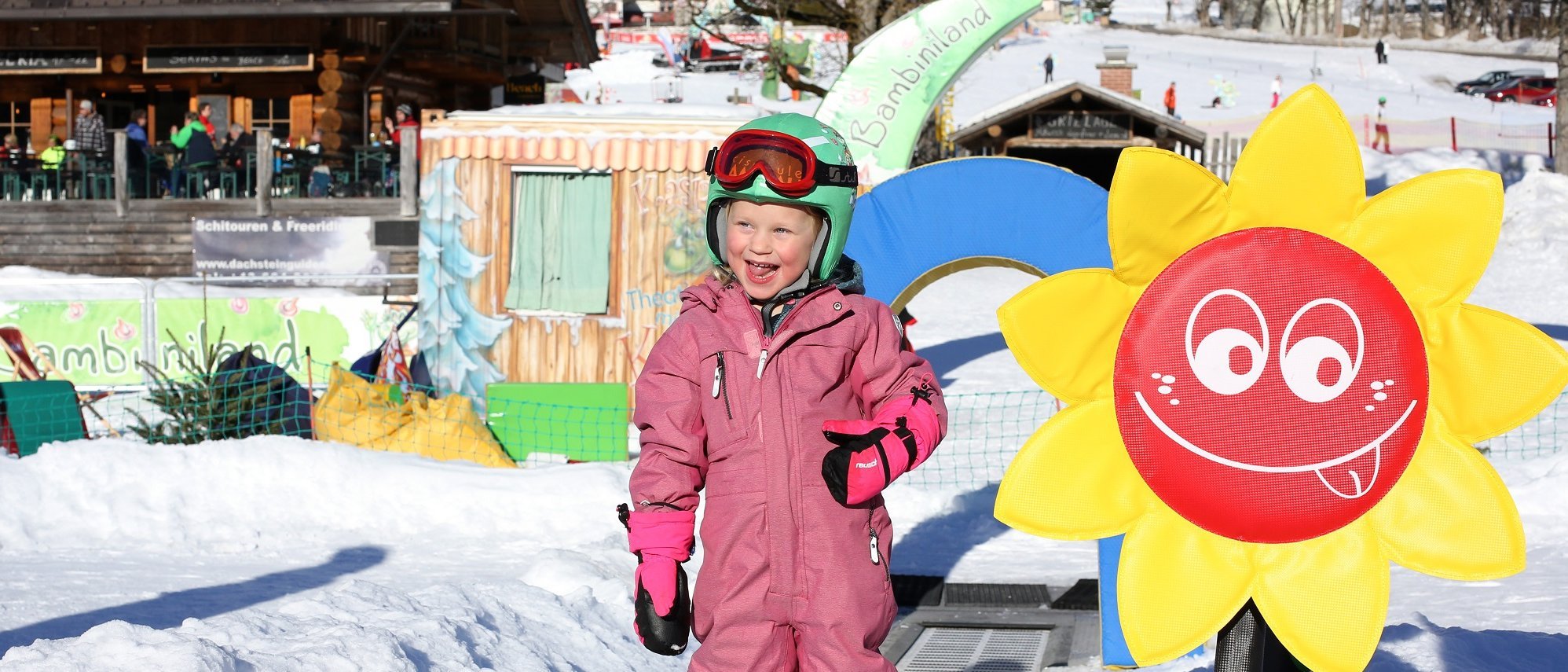 Winterspaß für Kinder Ein lächelndes Kind in einem rosa Skianzug steht auf einer schneebedeckten Piste mit einer bunten Sonnendekoration.