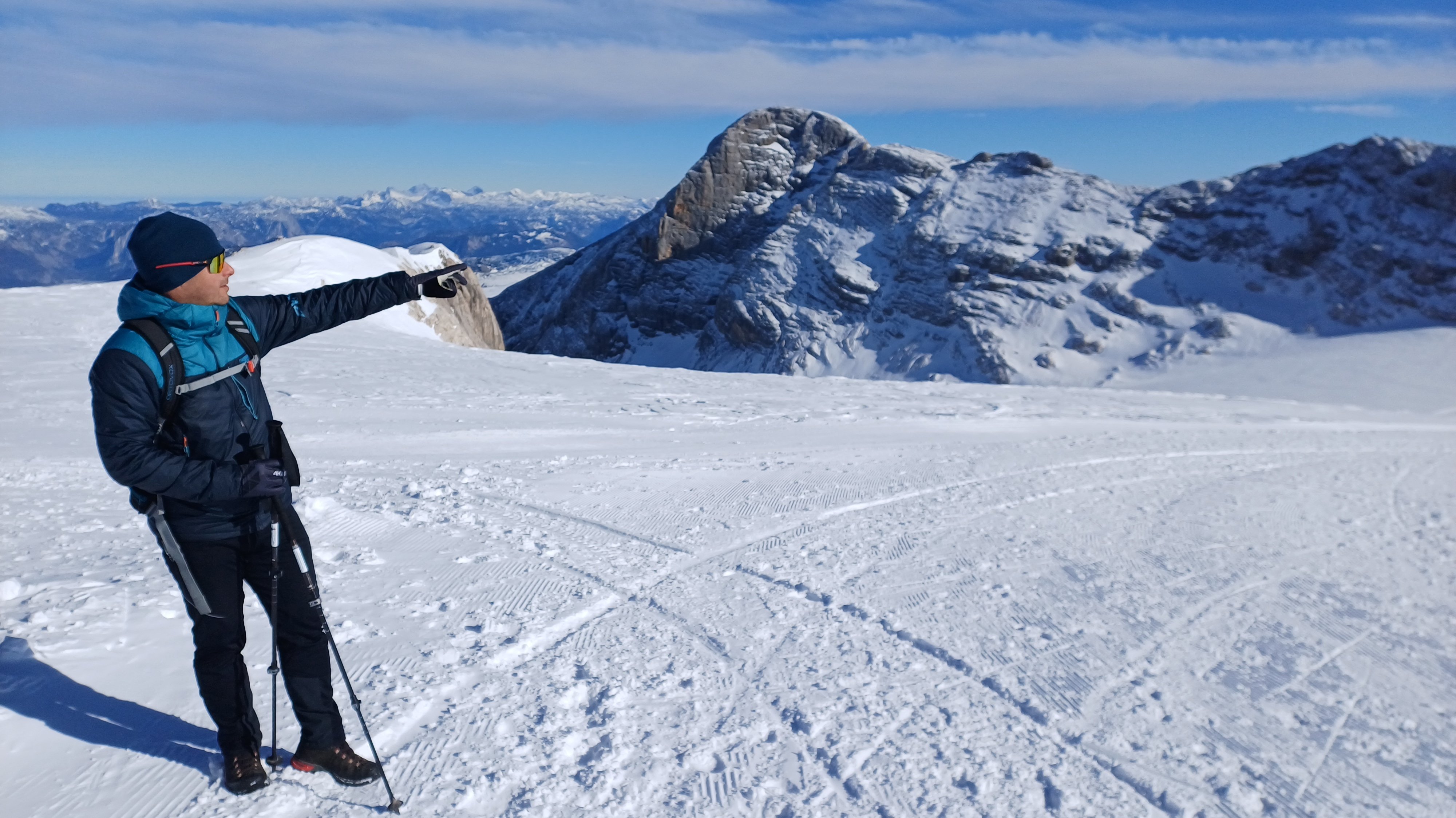 Abenteuer in verschneiter Berglandschaft Eine Person steht im Schnee und zeigt auf einen entfernten Gipfel unter klarem Himmel.