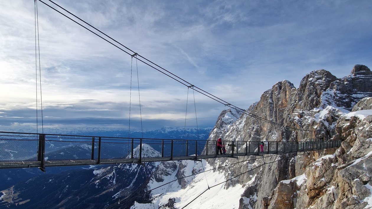 Schwebende Brücke im winterlichen Bergpanorama Suspension bridge in a snowy mountain landscape with hikers and rocky cliffs in background