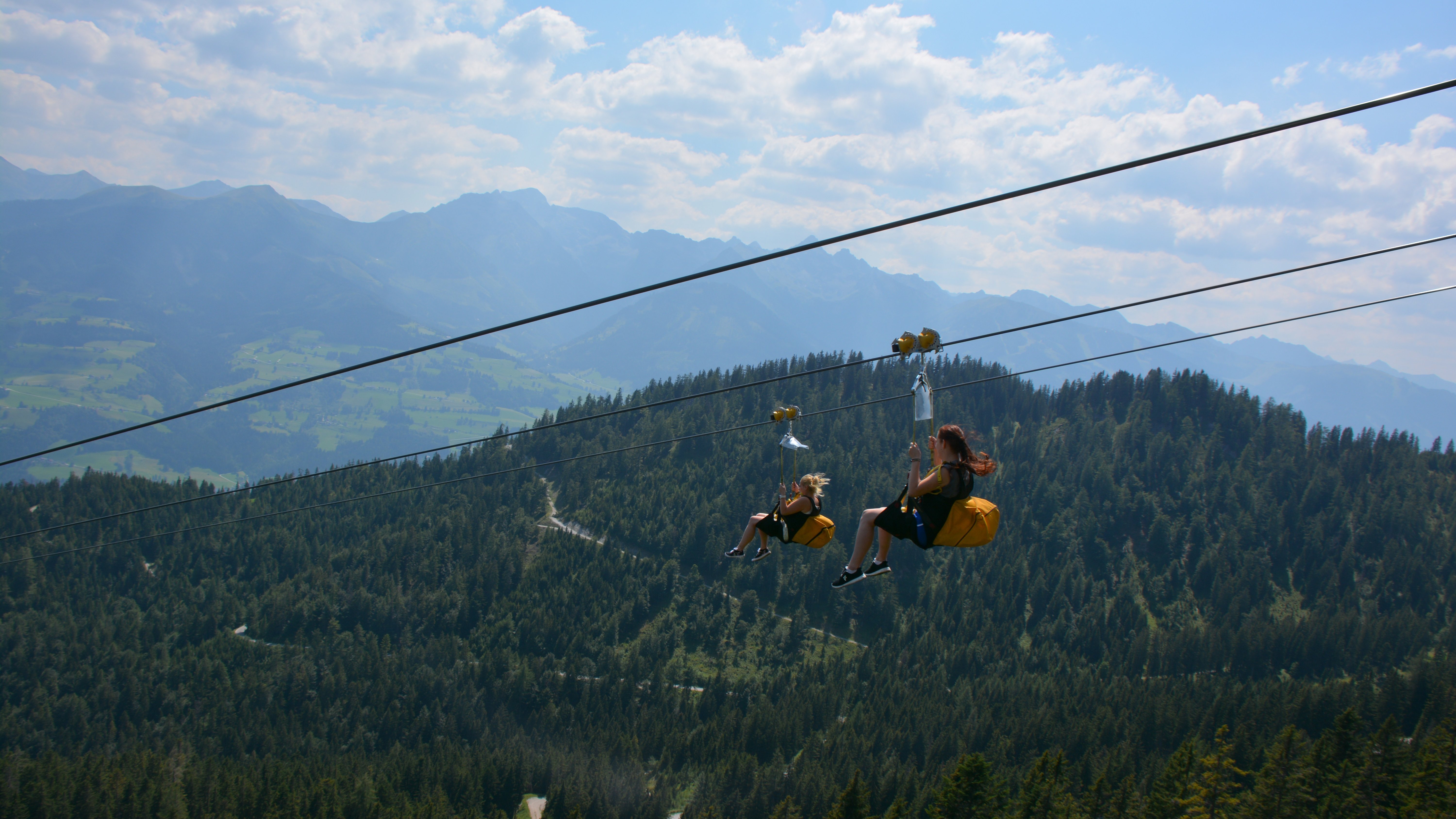Abenteuer auf der Zipline über den Bergen Zwei Personen fahren mit der Zipline über eine Berglandschaft mit Bäumen und Hügeln.