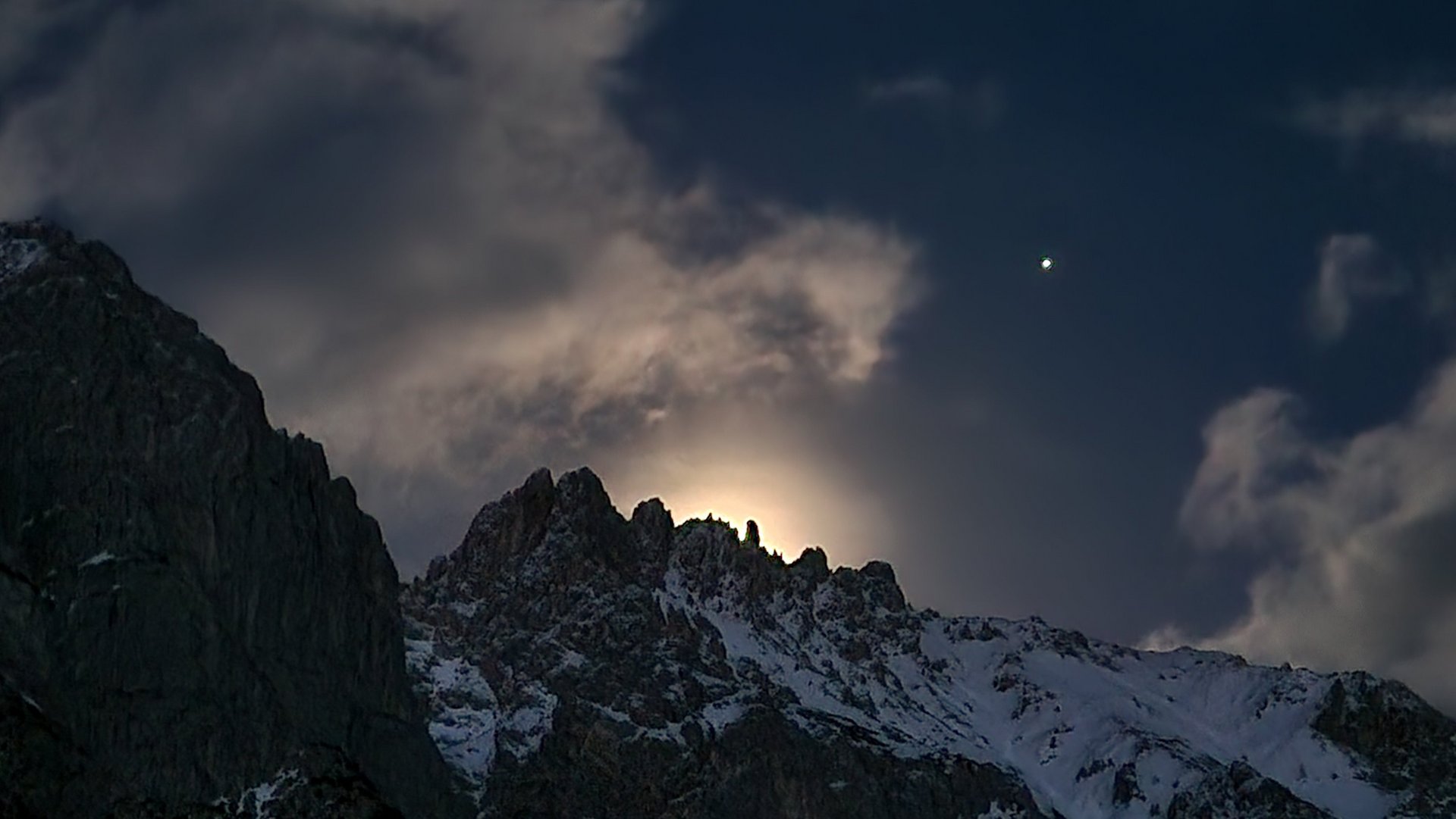 Berglandschaft bei Nacht mit sichtbarem Himmelskörper und Wolken im Hintergrund.