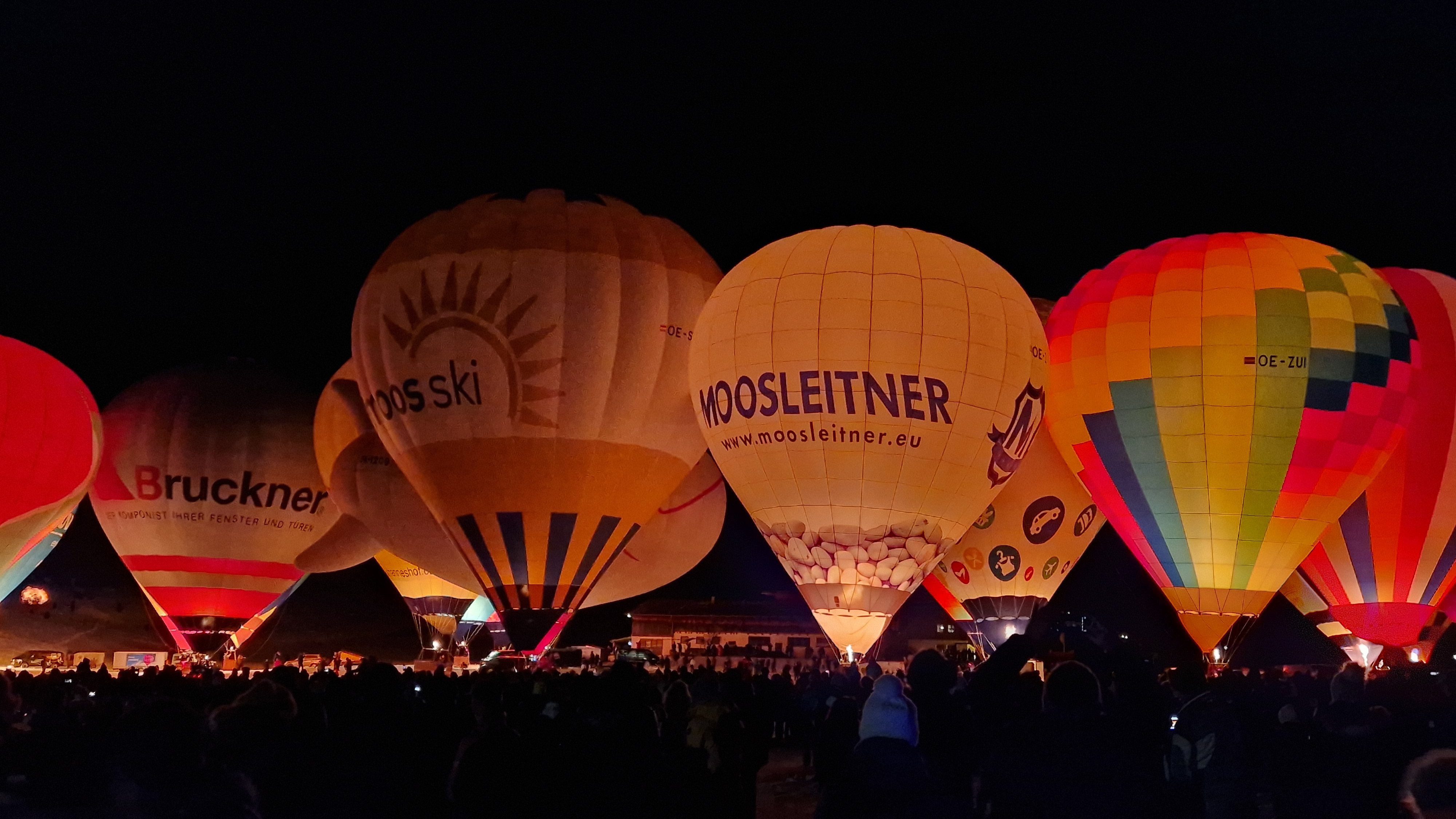 Illuminierte Ballons im Nachtglanz Mehrere Heißluftballons leuchten in der Nacht mit Menschenmenge darunter, bei einem Festival.