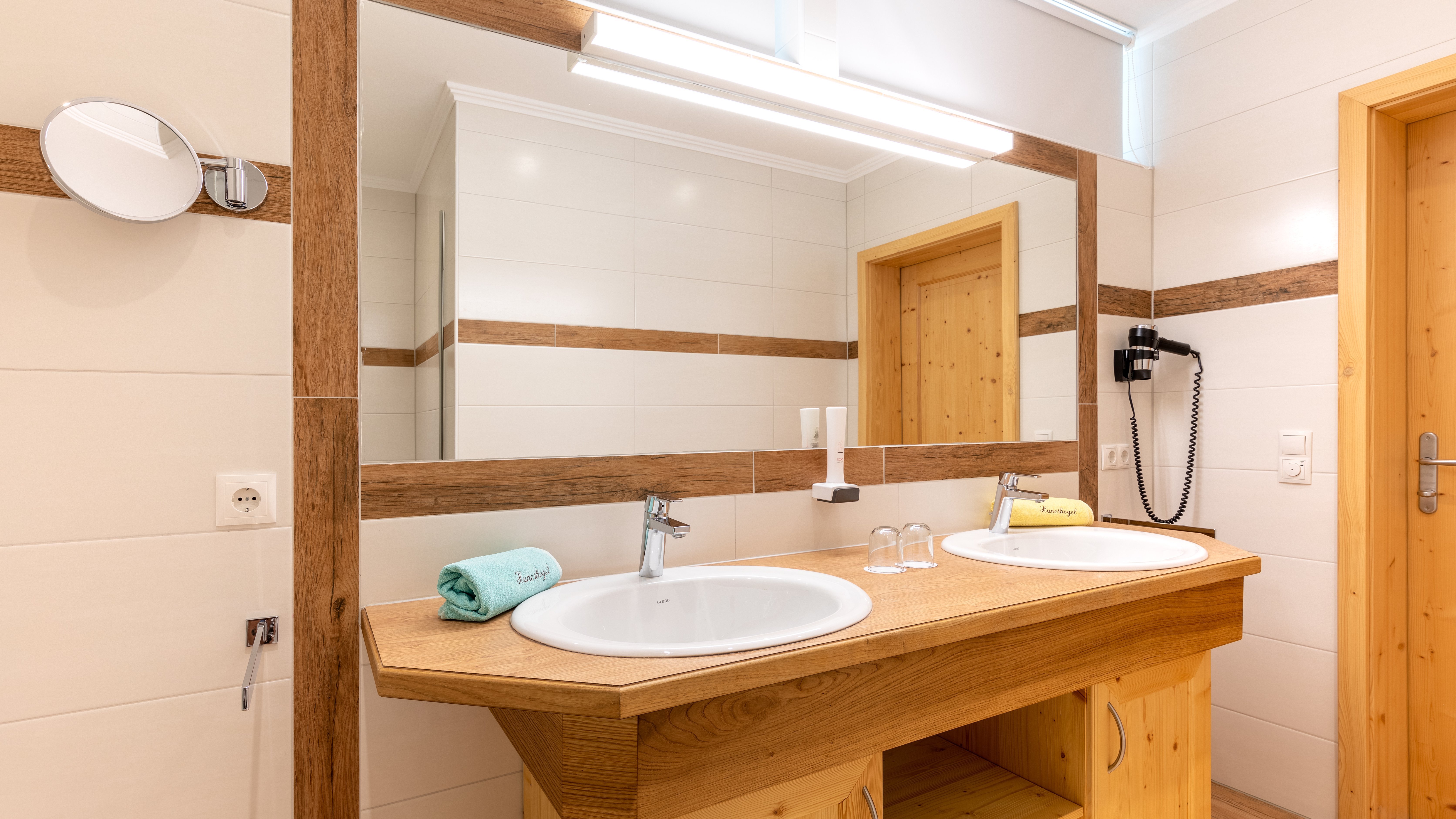Badezimmer mit Doppelwaschbecken und modernem Spiegel, Holzdetails sichtbar und stilvolle Beleuchtung.
