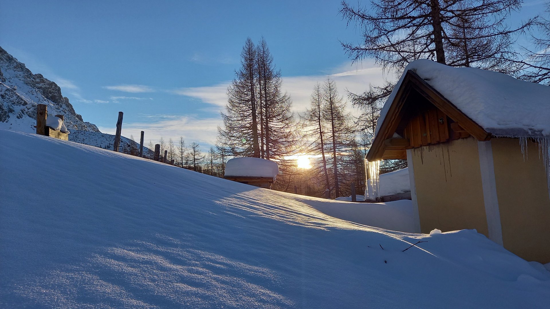 Schneebedeckte Landschaft mit einer Hütte und schneebedeckten Bäumen vor Bergen bei Sonnenuntergang