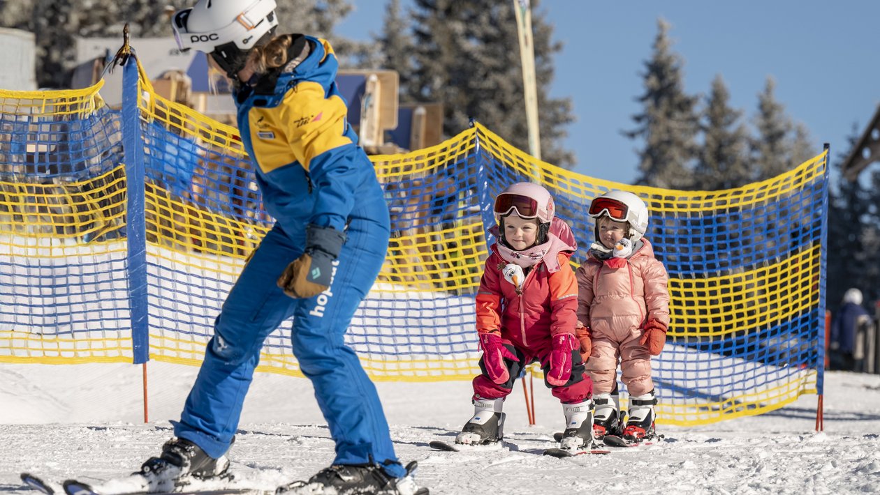 Skikurs für Kinder in Ramsau Skiinstruktor lehrt zwei Kinder das Skifahren auf einer schneebedeckten Piste mit Sicherheitsbarrieren im Hintergrund.