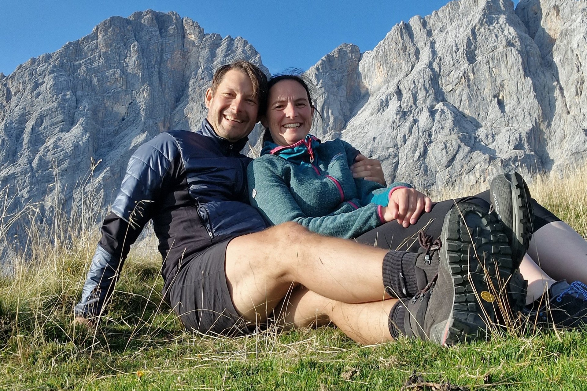 Ein Paar sitzt auf einer Wiese in den Bergen unter einem blauen Himmel und genießt die Natur.