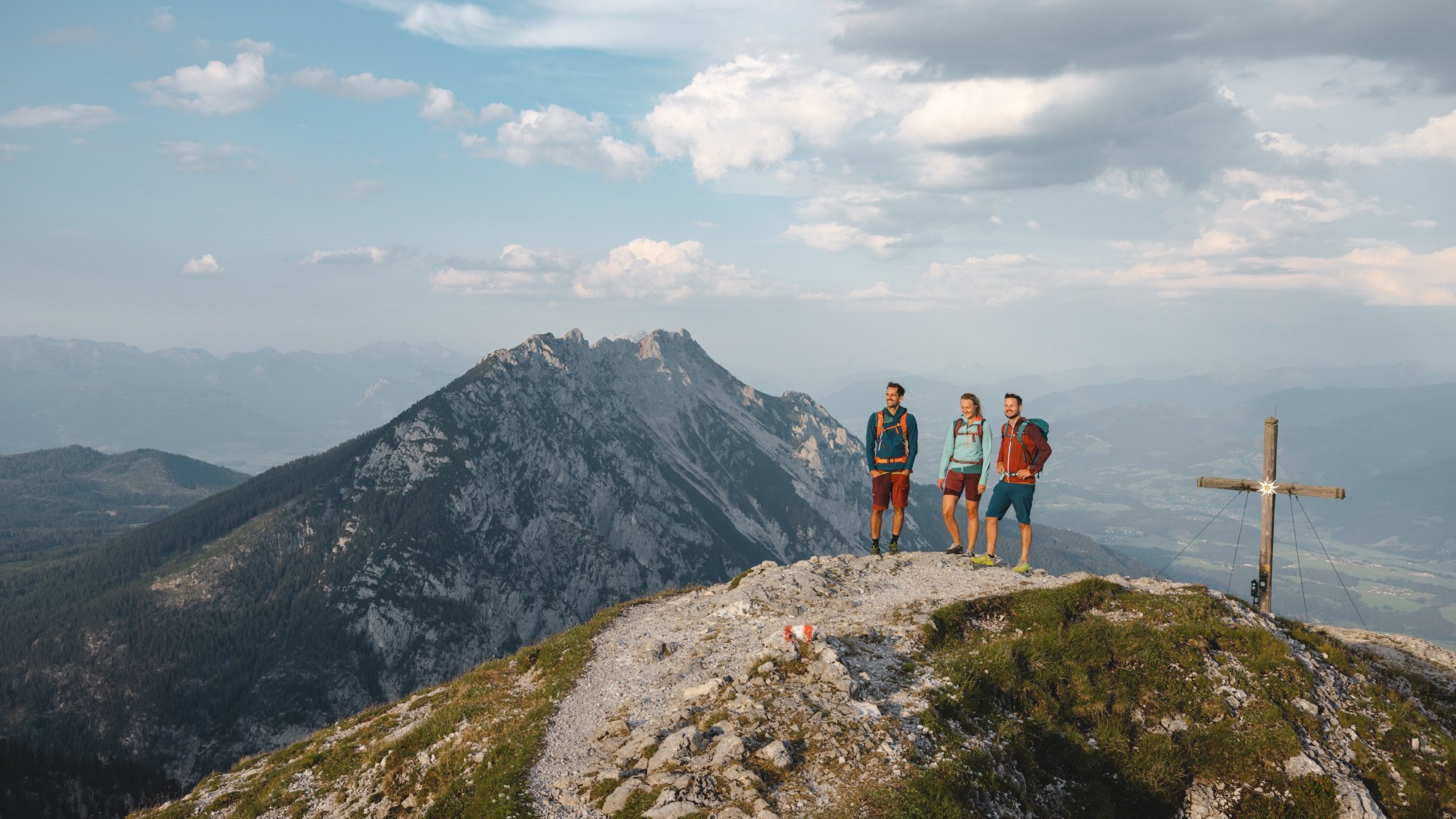 Hiking Adventure on the Mountain Peak Drei Wanderer stehen auf einem felsigen Berggipfel mit einem Kreuz im Vordergrund und Blick auf die Landschaft.
