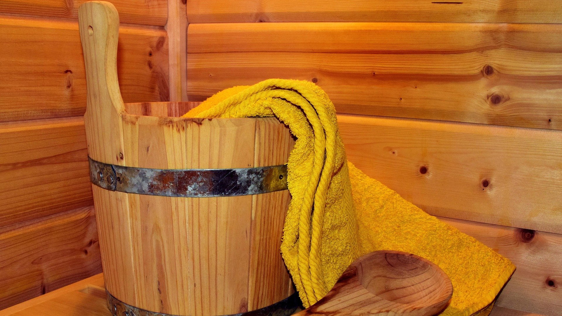 Holzbucket mit Metallband und gelbem Handtuch, Holzlöffel daneben vor Holzwand bei Sauna