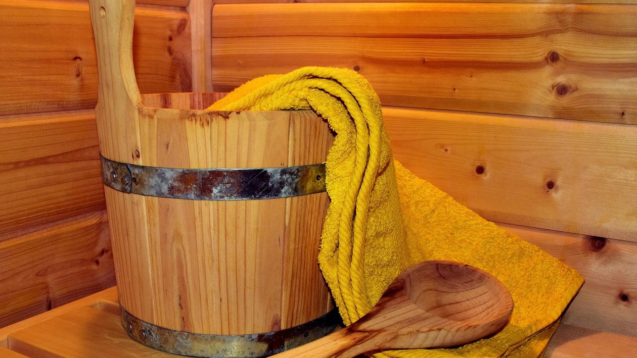 Wohlfühlmomente in der Sauna Holzbucket mit Metallband und gelbem Handtuch, Holzlöffel daneben vor Holzwand bei Sauna