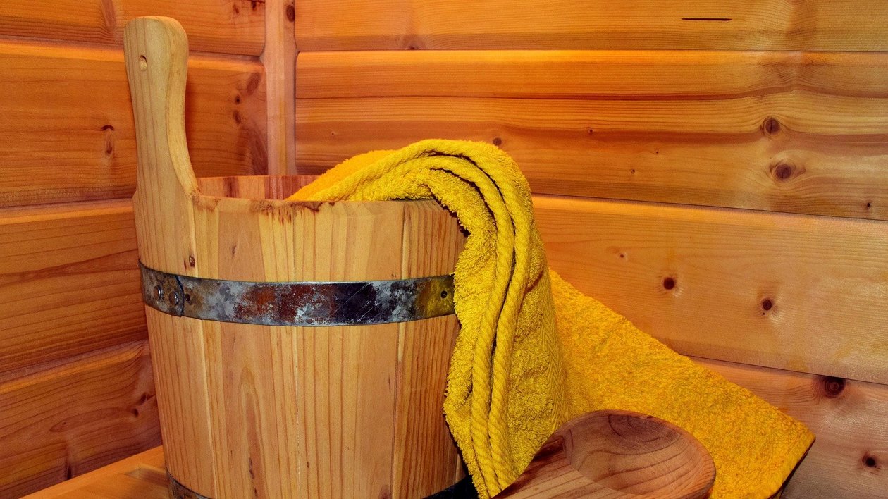 Wohlfühlmomente in der Sauna Holzbucket mit Metallband und gelbem Handtuch, Holzlöffel daneben vor Holzwand bei Sauna