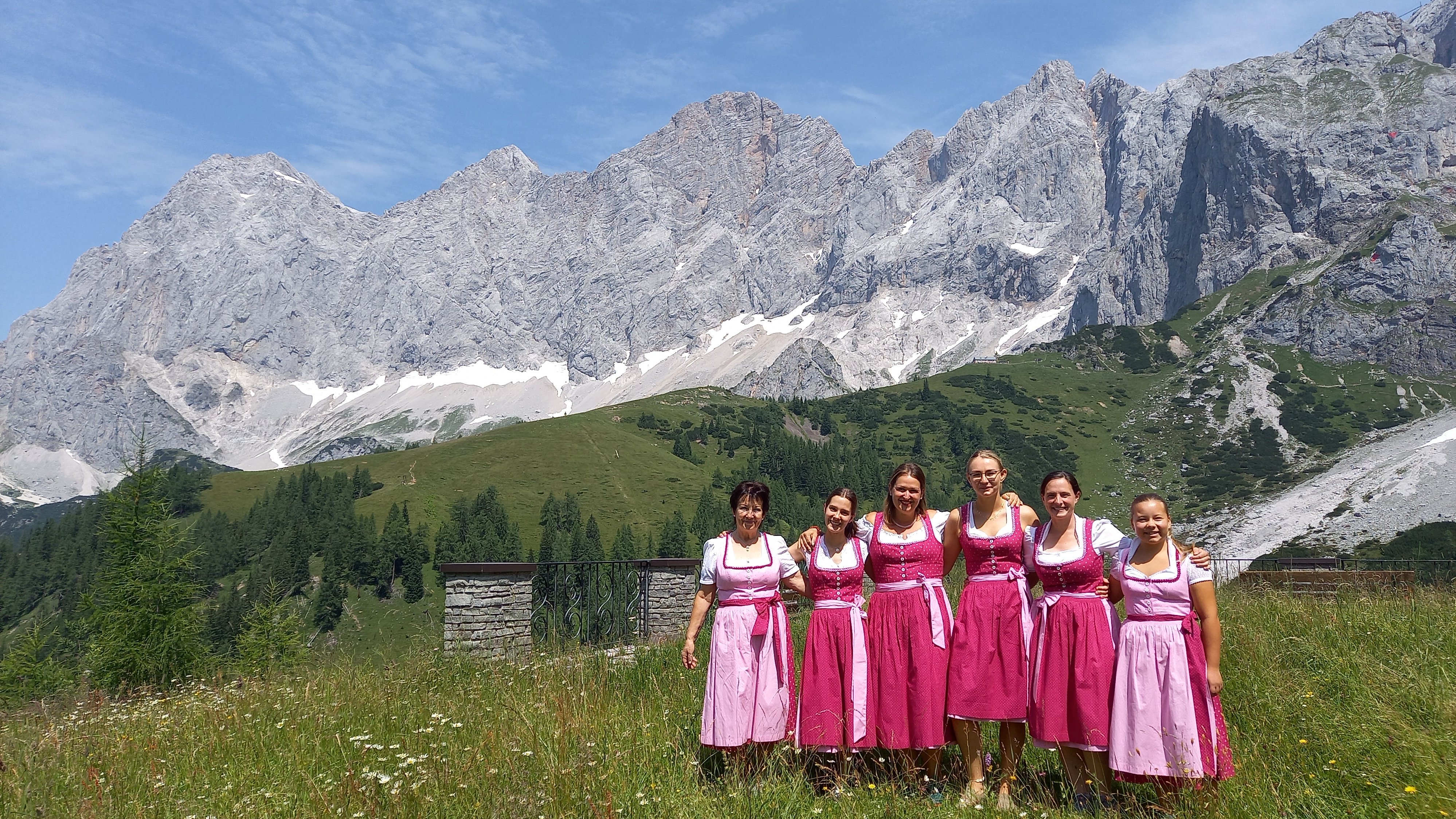 Traditionelle Trachten im Alpenpanorama Gruppe von sechs Frauen in traditionellen Kleidern auf einer Wiese mit Berglandschaft im Hintergrund