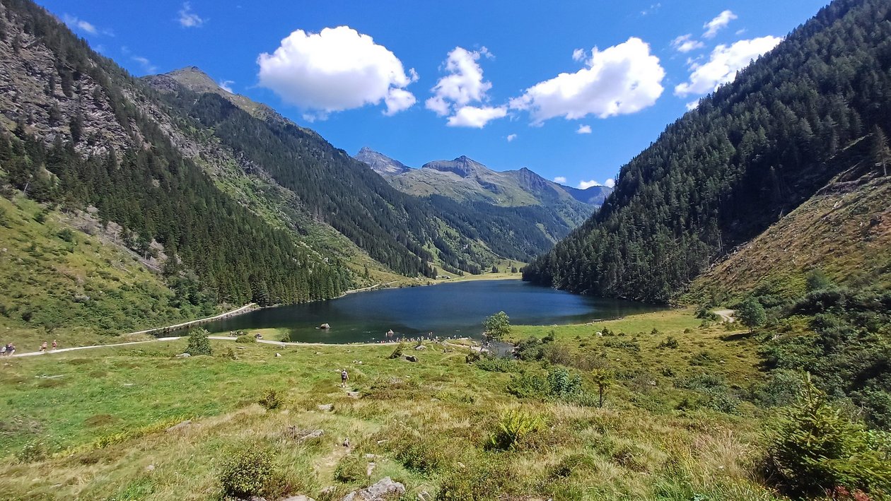 Idyllischer Bergsee inmitten grüner Hügel Ruhiger Bergsee umgeben von grünen Hügeln und Bergen unter klarem blauen Himmel mit Wolken