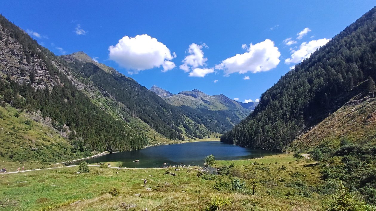 Idyllischer Bergsee inmitten grüner Hügel Ruhiger Bergsee umgeben von grünen Hügeln und Bergen unter klarem blauen Himmel mit Wolken