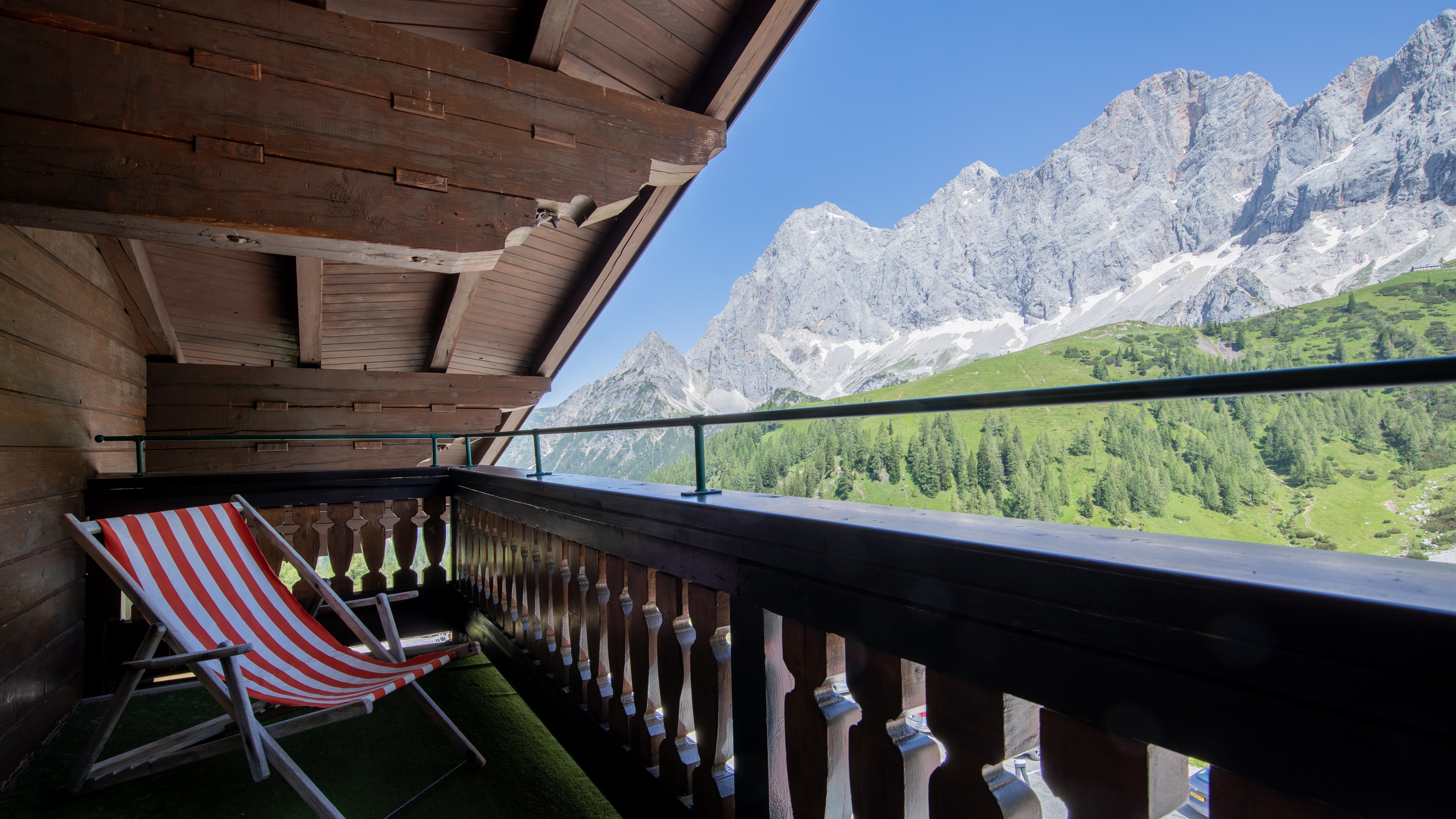 Balkon mit Holzarchitektur und Stuhl, Blick auf Berge mit Schnee und grünen Wiesen