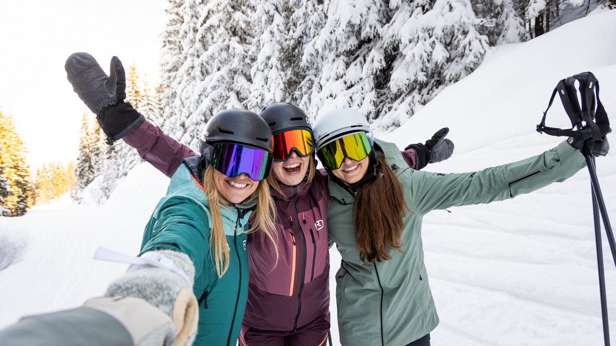 Drei Frauen stehen lächelnd im Schnee und posieren beim Skifahren in einer winterlichen Landschaft.