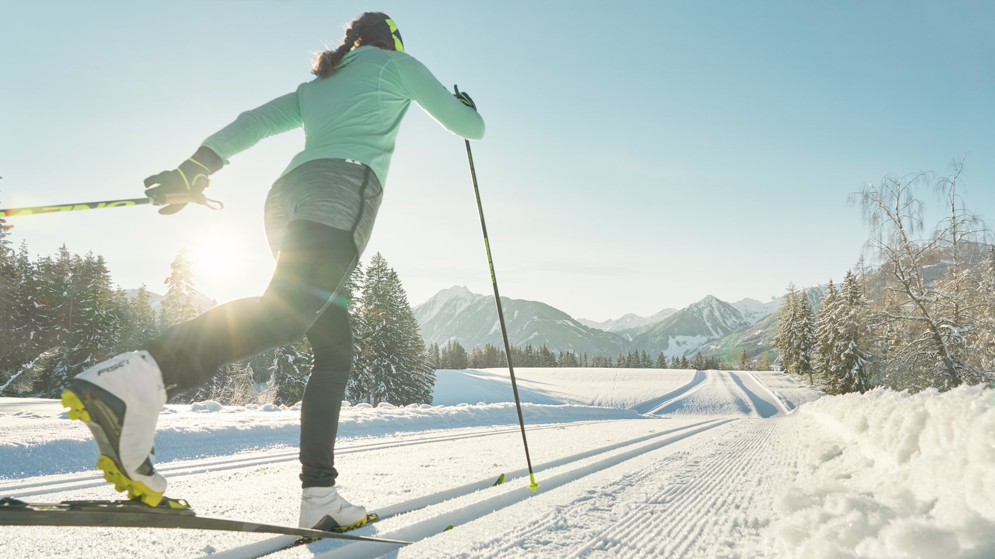 Langlaufen in der Winterlandschaft Person beim Langlaufen auf einem verschneiten Weg mit Bergen und Bäumen im Hintergrund.