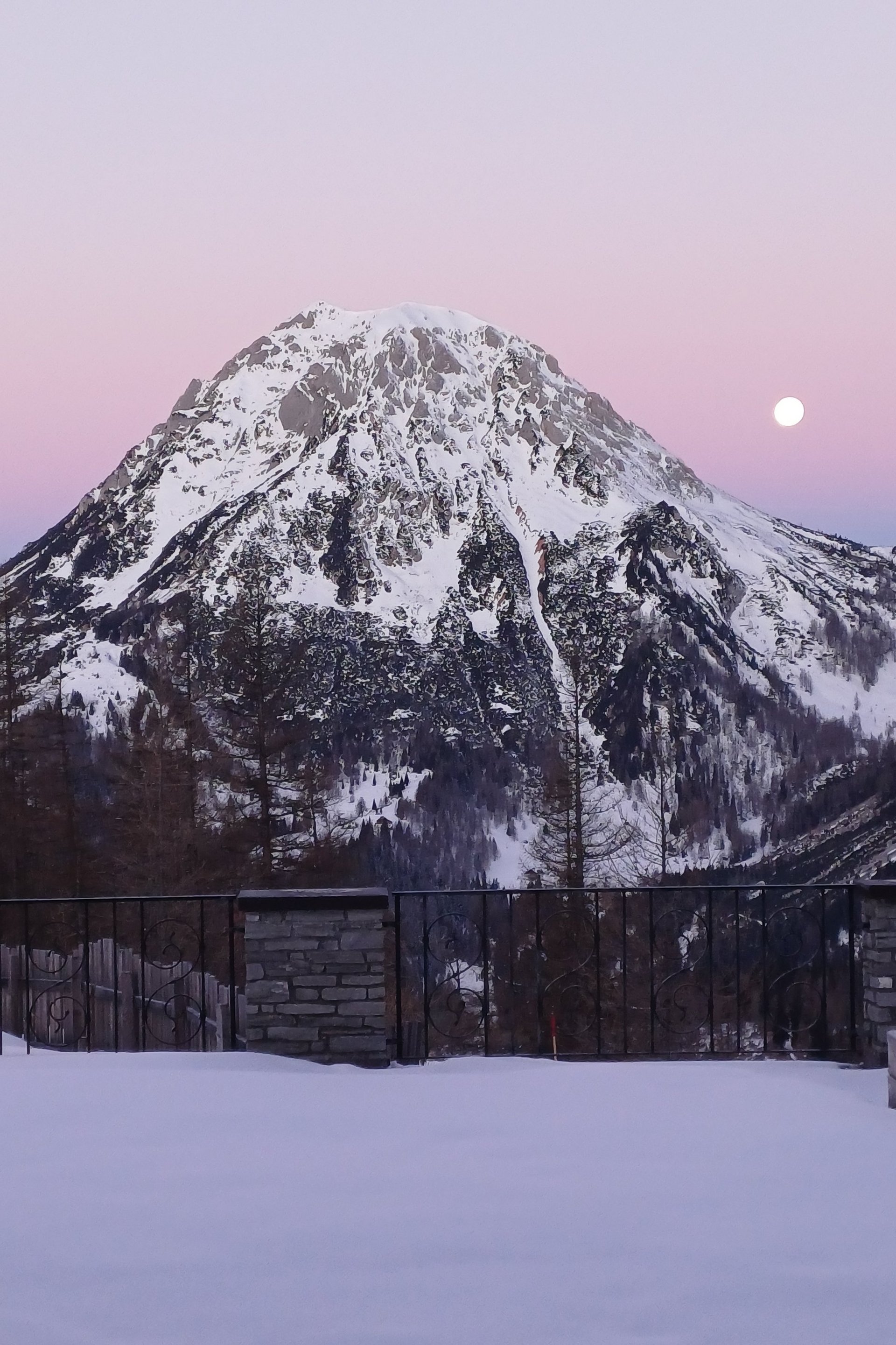 Verschneiter Berg mit aufgehender Mondsichel vor pastellfarbenem Himmel bei Dämmerung