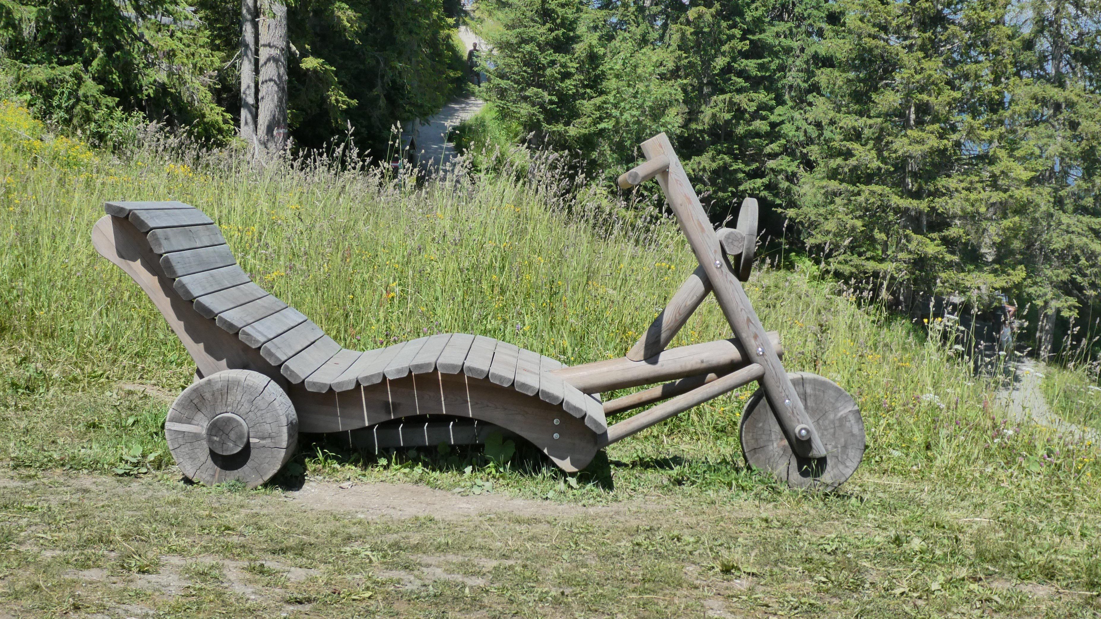 Holzskulptur in der Natur Holzskulptur in Form eines Fahrrads in einer Wiese umgeben von Bäumen und Grünflächen.