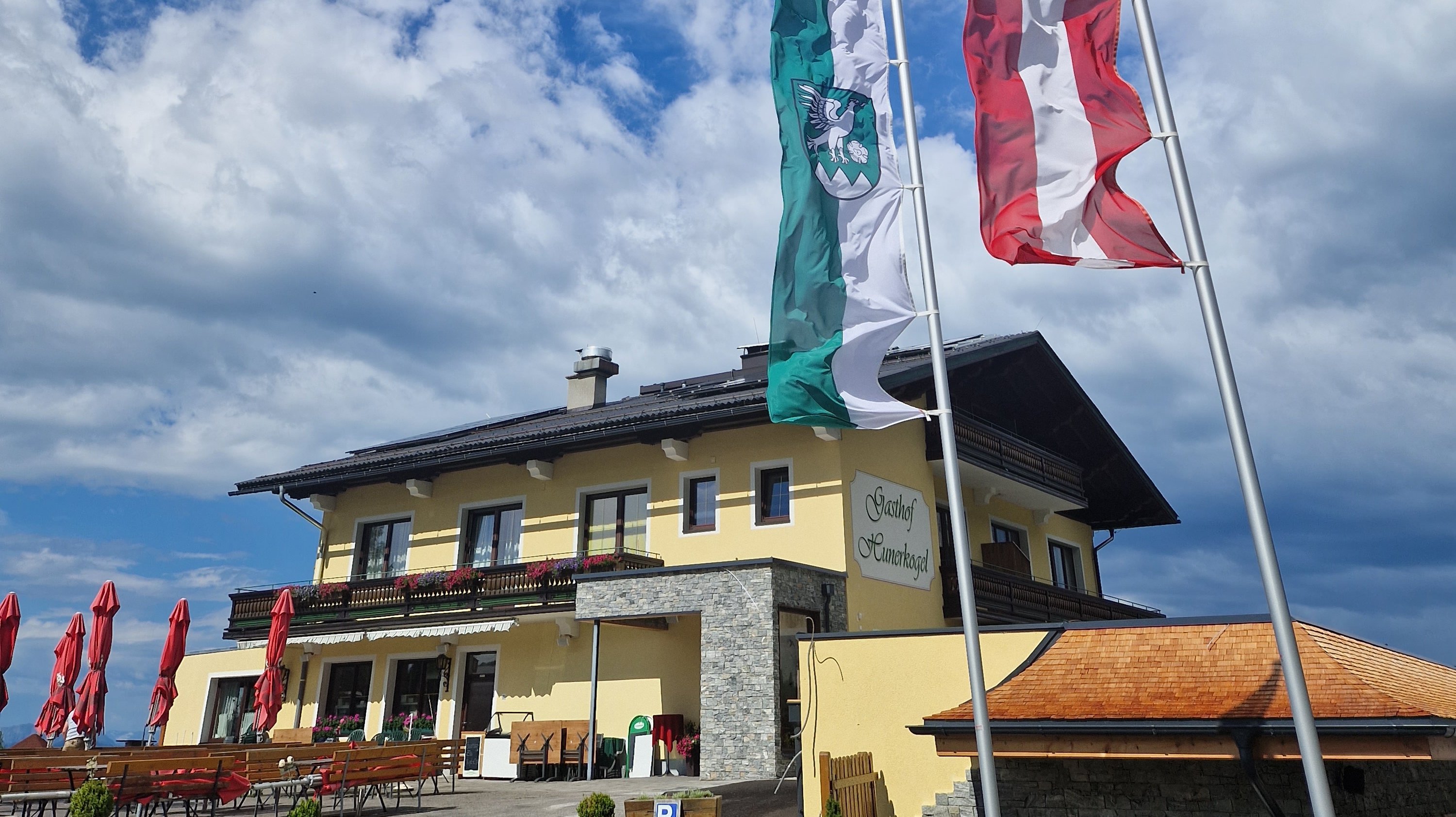 Berggasthof mit Blick auf die Berge Restaurantgebäude mit zwei Fahnen vor blauem Himmel und Wolken im Hintergrund. Außensitzplätze sind sichtbar.