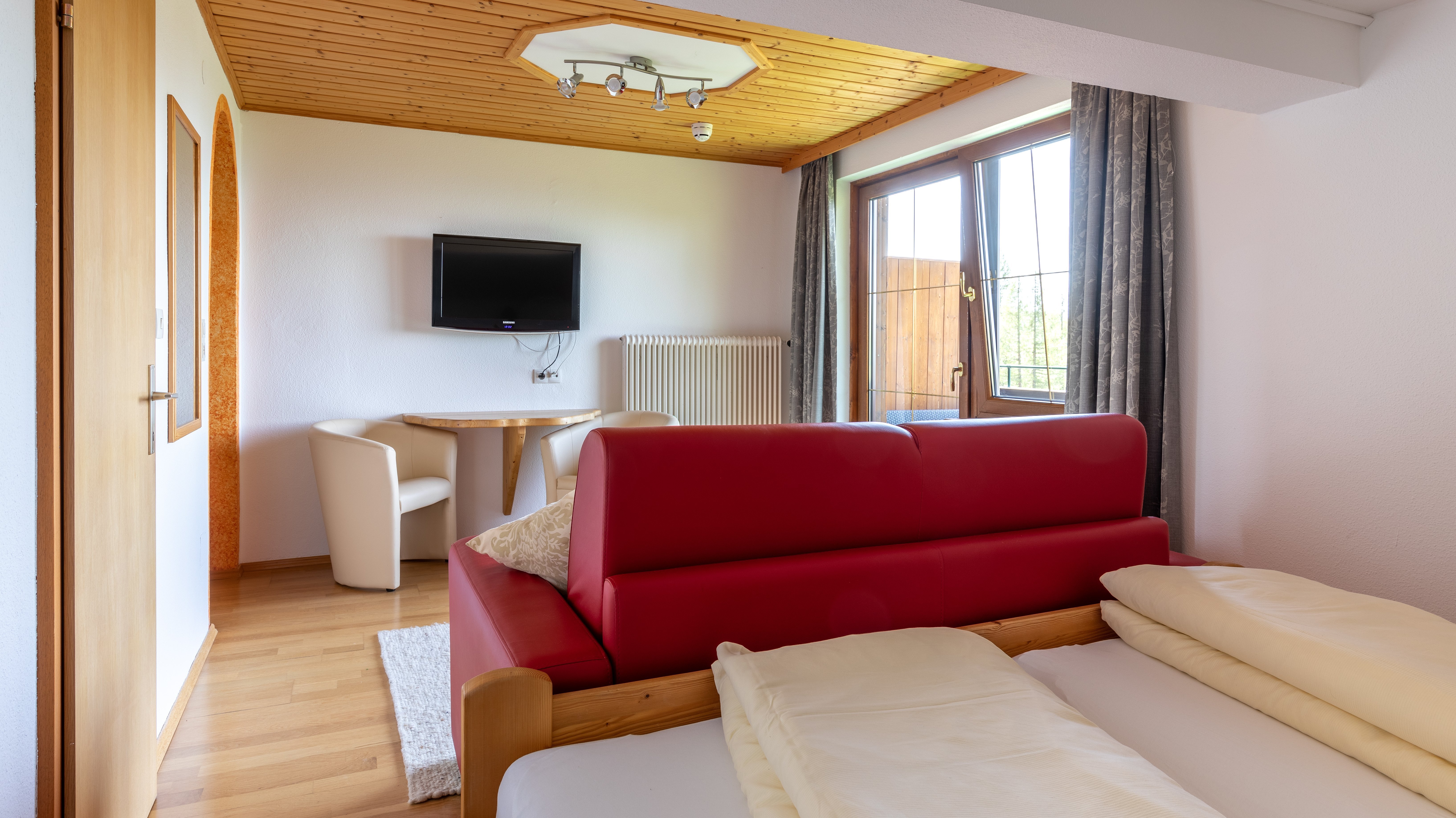 Hotelzimmer mit rotem Sofa, Holztisch und Fernseher an der Wand, große Fenster bieten Tageslicht.
