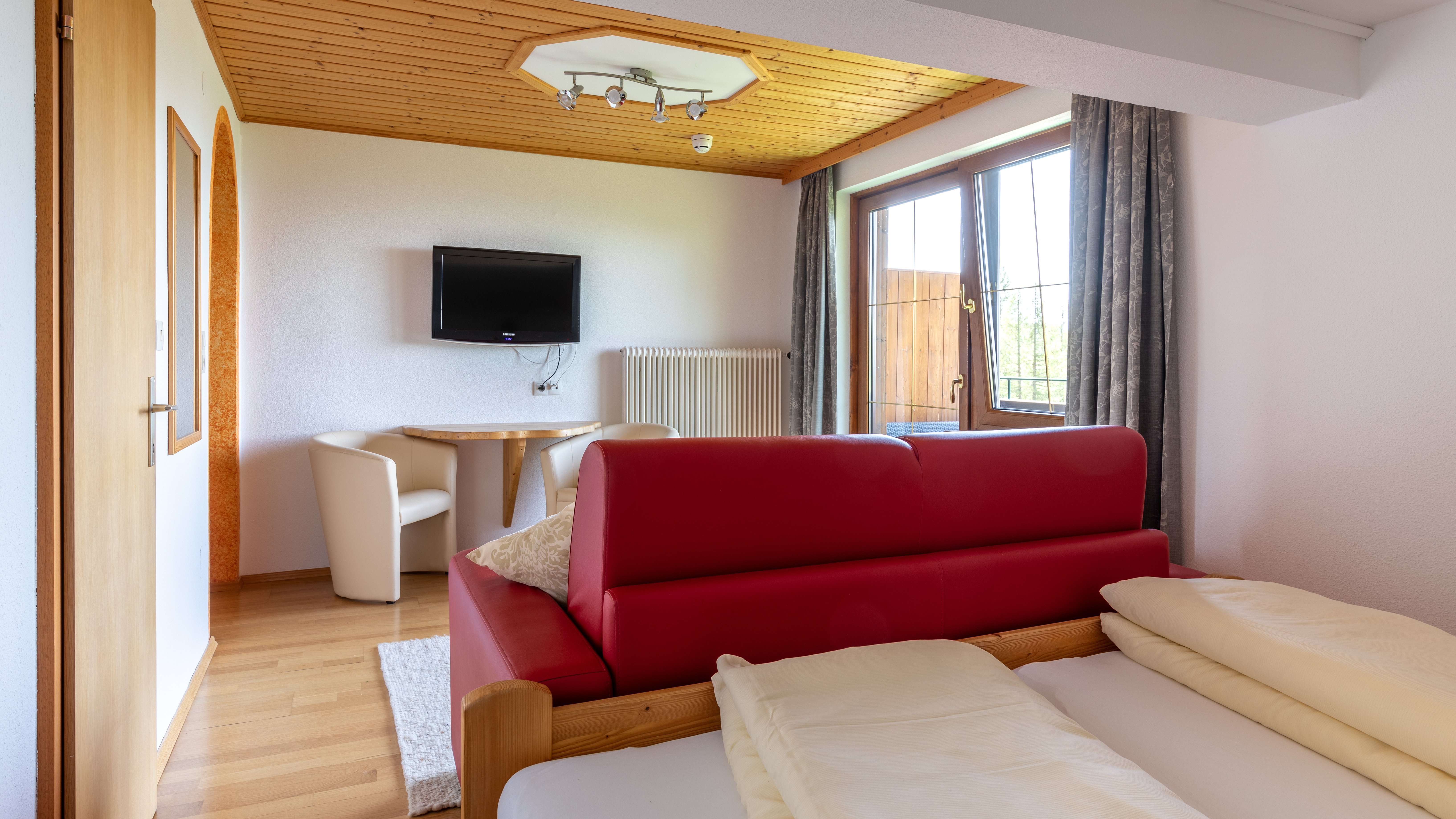 Hotelzimmer mit rotem Sofa, Holztisch und Fernseher an der Wand, große Fenster bieten Tageslicht.