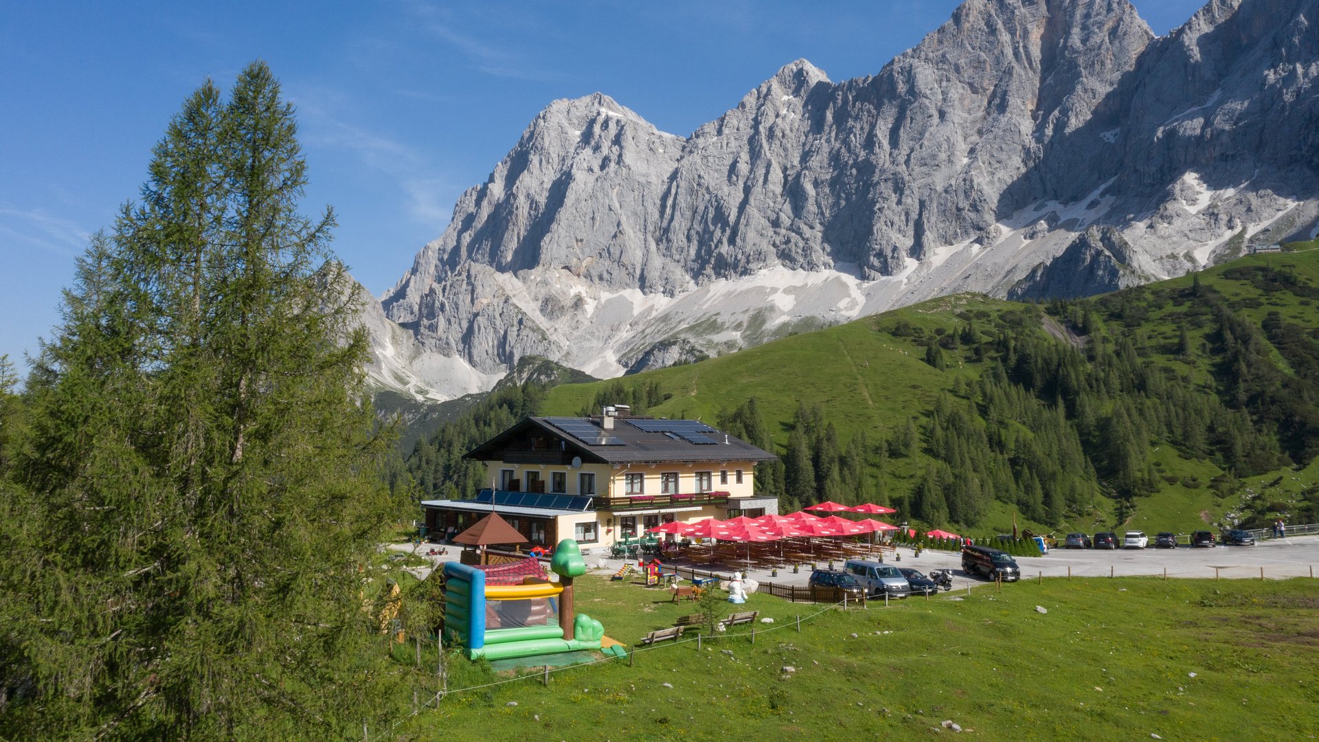 Blick auf einen Gasthof mit Spielplatz und Berglandschaft im Hintergrund und blauen Himmel