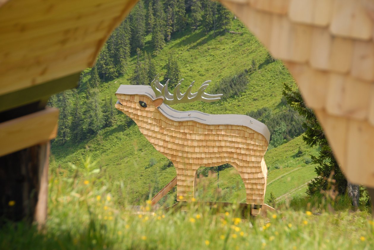 Holzskulptur im Bergpanorama Eine große Holzskulptur eines Rehs steht in einer grünen Berglandschaft, umgeben von Holzstrukturen.