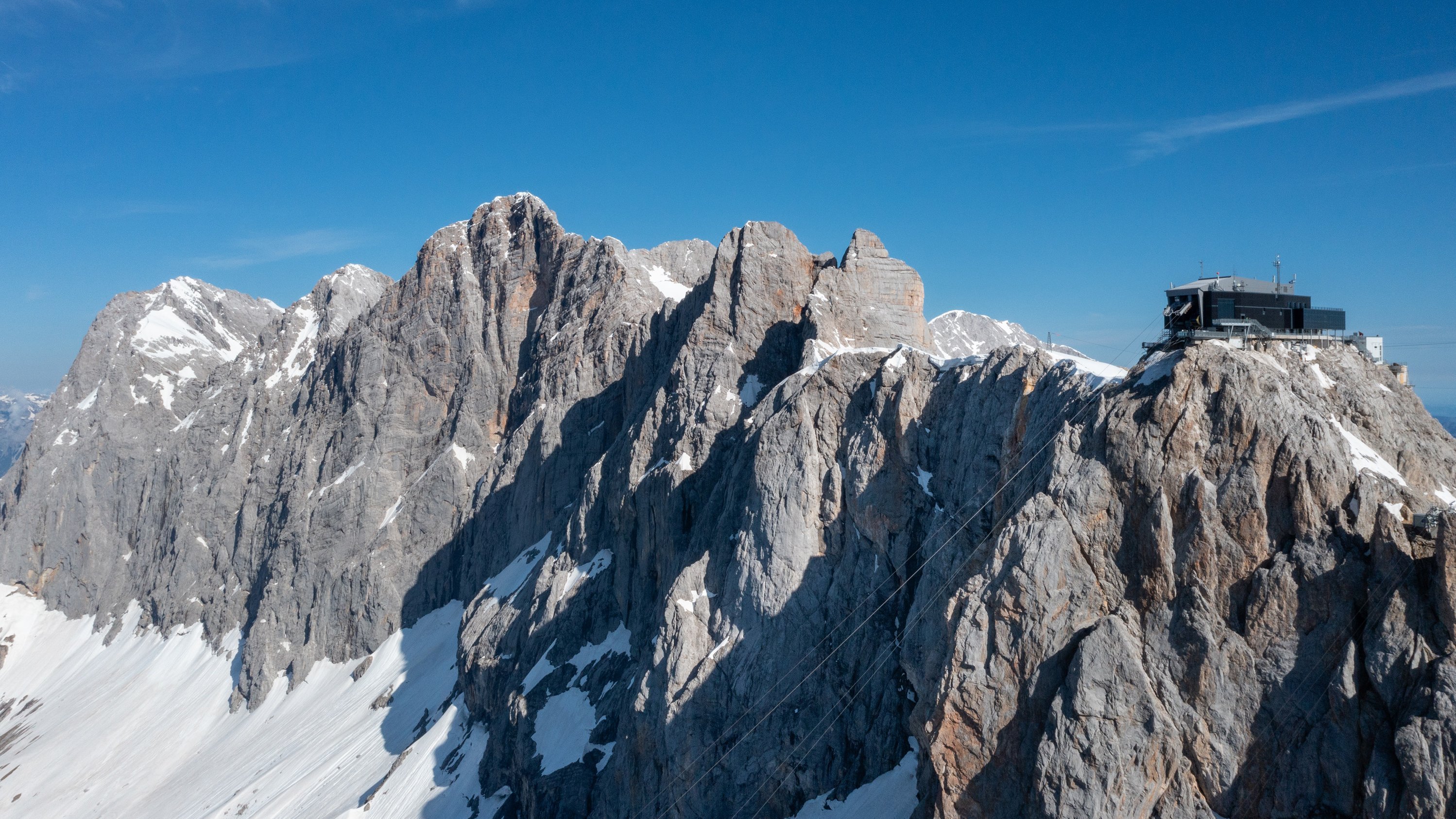Luftaufnahme einer gebirgigen Landschaft mit schneebedeckten Gipfeln und einem Gebäude auf einer Anhöhe.