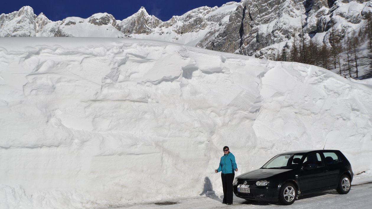 Winterlandschaft mit Fahrzeug und Frau Eine Person steht neben einem geparkten Auto an einem großen Schneehaufen vor Bergen.