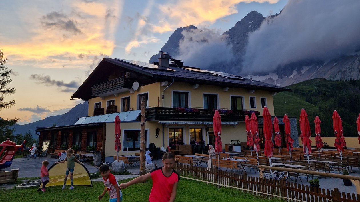 Familienfreuden am Berg Kinder spielen Fußball auf einer Wiese vor einem Berggasthof bei Sonnenuntergang mit dramatischen Wolken und Bergen im Hinter