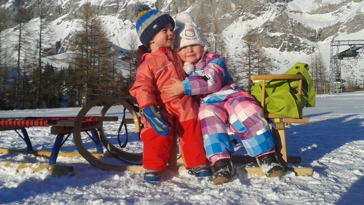 Winterspaß mit Kindern Zwei Kinder sitzen auf einer Holzbank in einer verschneiten Landschaft mit Bergen im Hintergrund.
