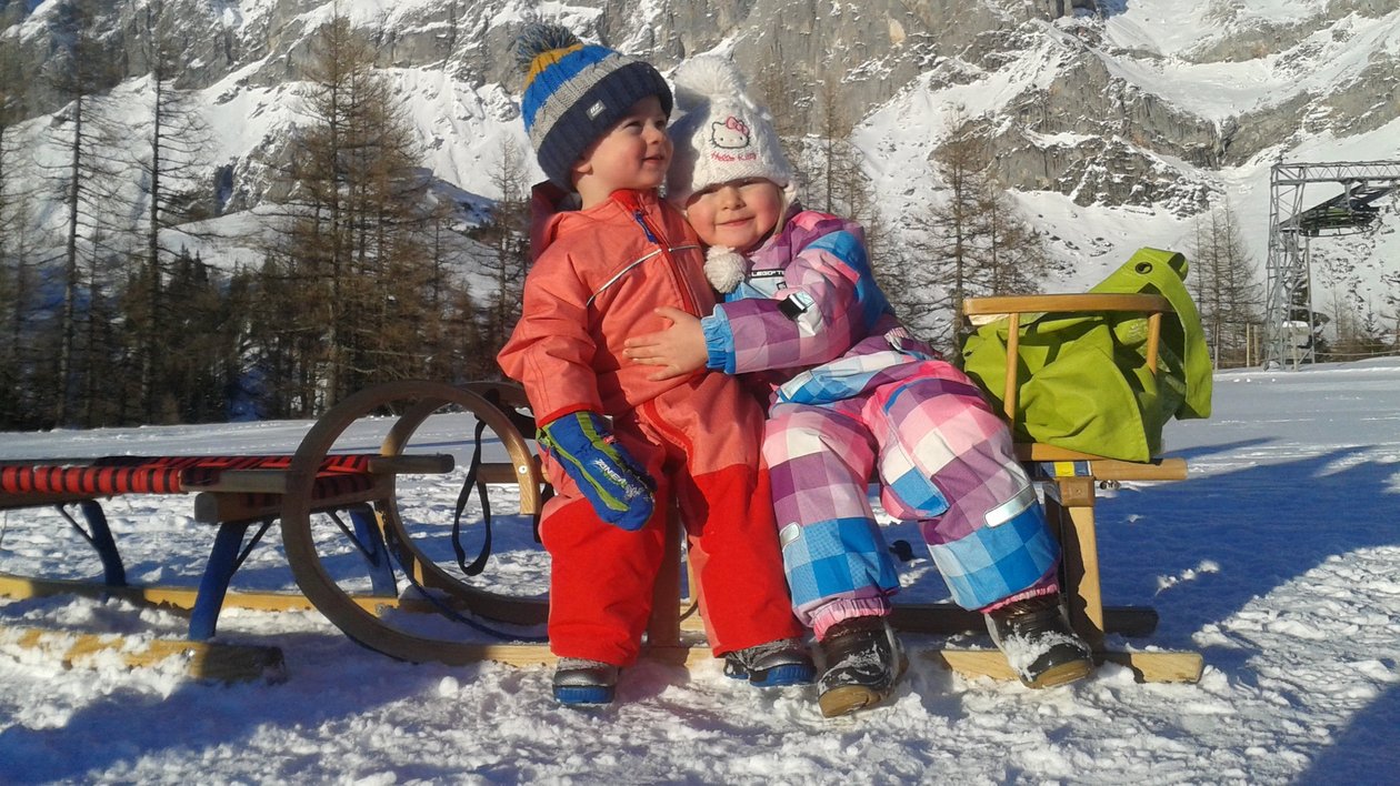 Winterspaß mit Kindern Zwei Kinder sitzen auf einer Holzbank in einer verschneiten Landschaft mit Bergen im Hintergrund.