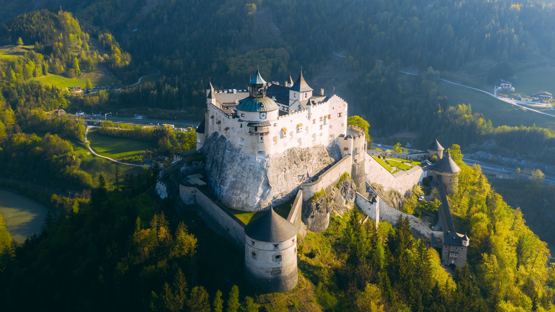 Luftaufnahme einer großen Burg auf einem Felsen, umgeben von Wäldern und Bergen.