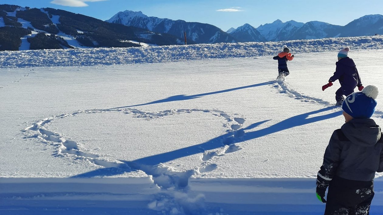 Kinder im Winterzauber Kinder spielen im Schnee und formen ein Herz, mit Bergen im Hintergrund sichtbar.