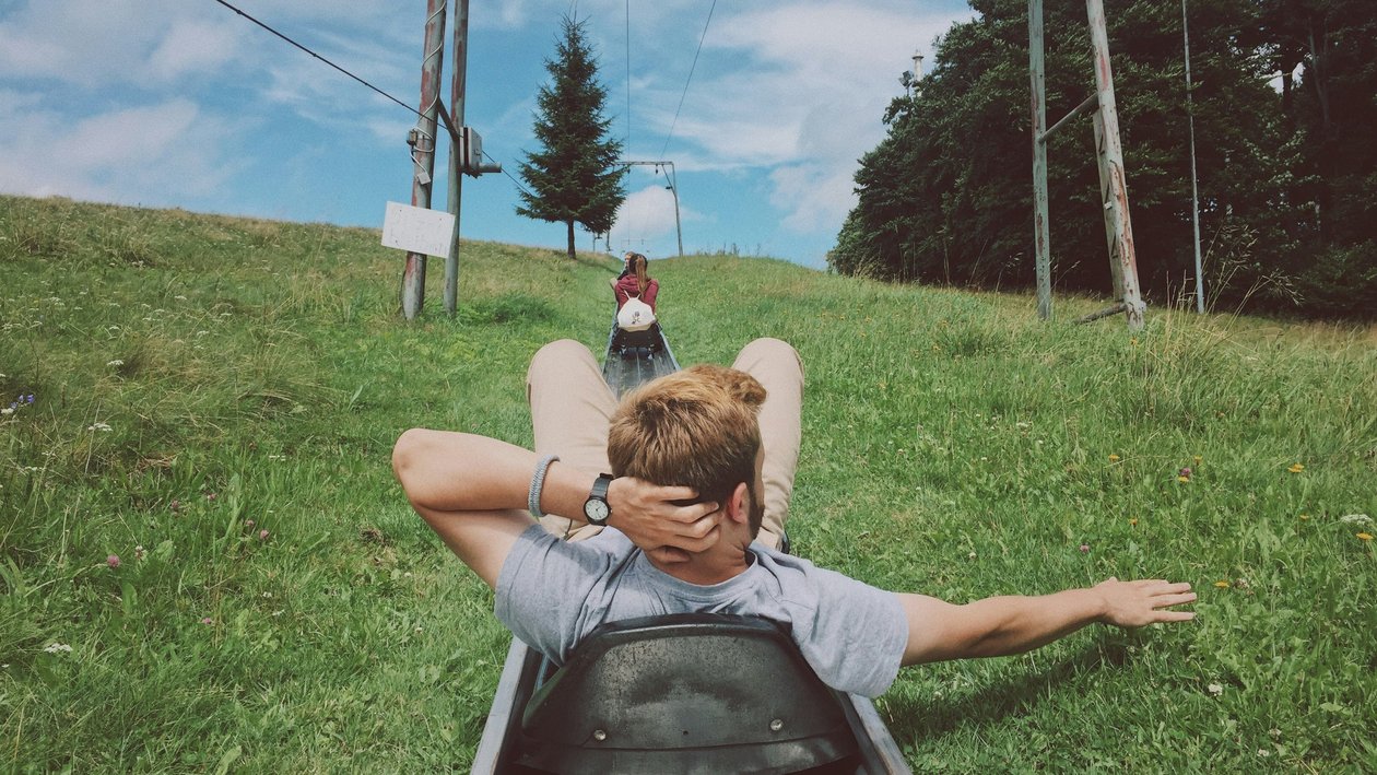 Person on a luge track descending a grassy slope with another rider in the background