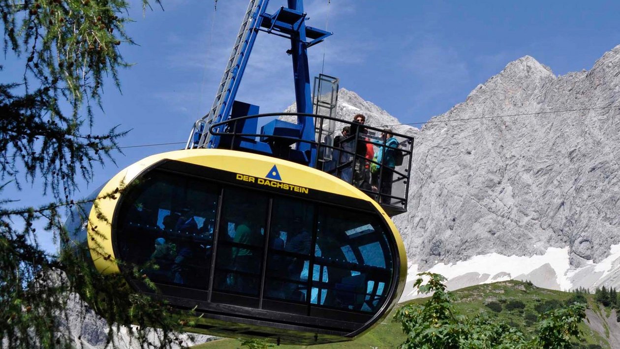 Seilbahnfahrten über den Dachstein Eine Seilbahn fährt über felsige Berggipfel mit grünen Wiesen im Hintergrund in den Alpen.