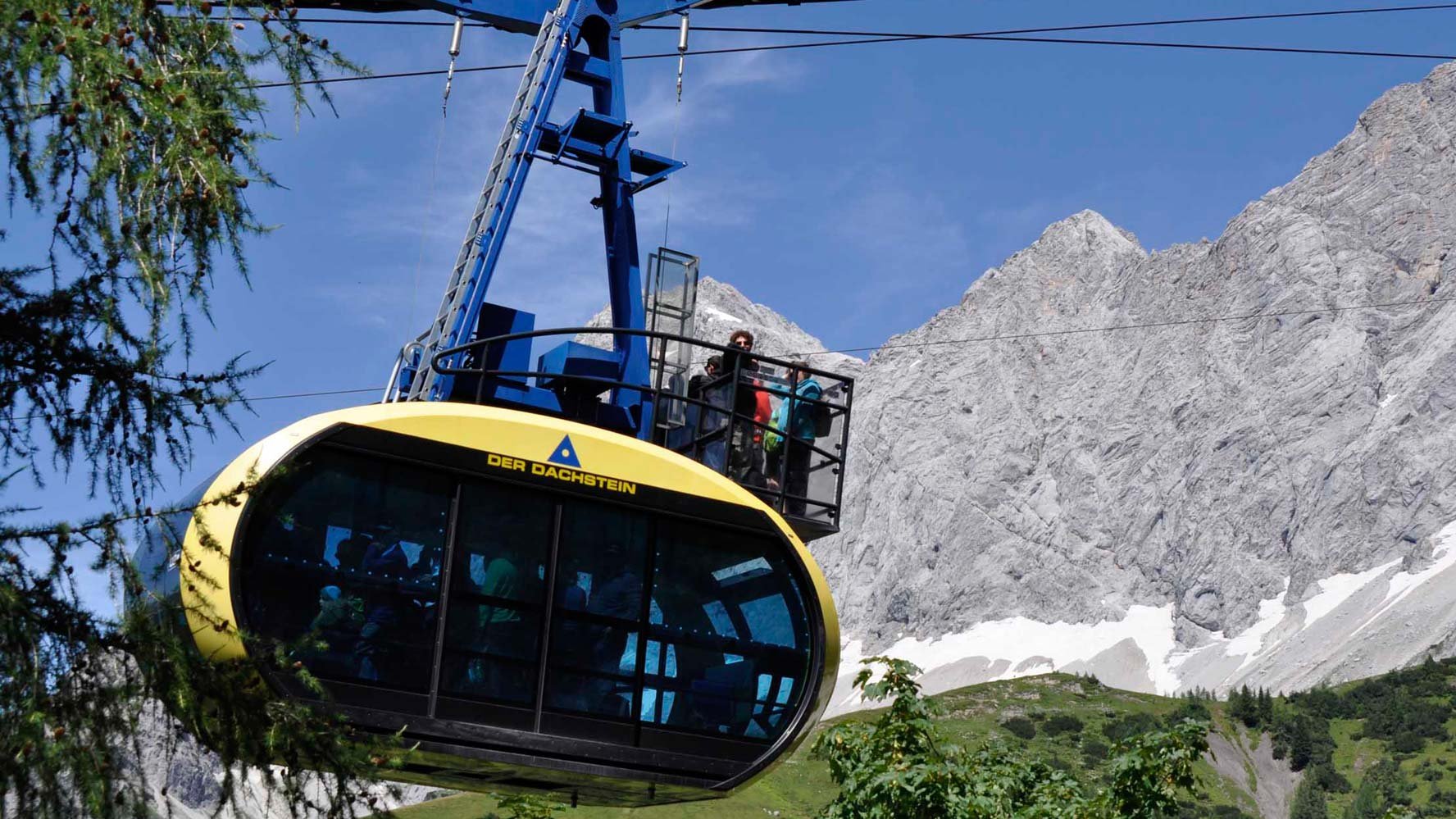 Eine Seilbahn fährt über felsige Berggipfel mit grünen Wiesen im Hintergrund in den Alpen.
