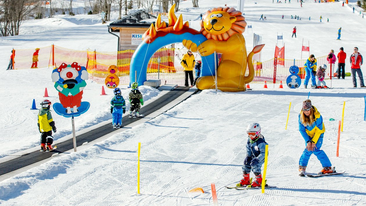Kinder im Skispaß Gruppe von Kindern beim Skifahren auf einer schneebedeckten Piste mit einem bunten Aufblaslöwen und Skilehrern im Hintergrund